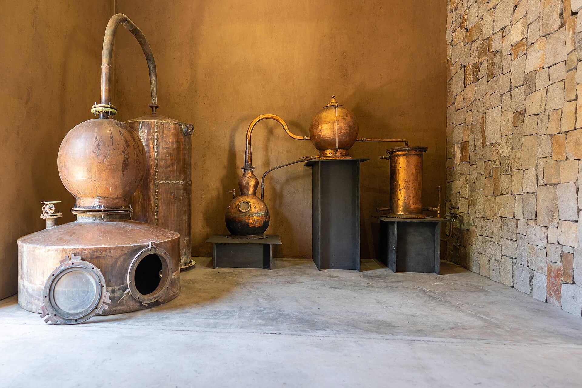Alambiques de cobre em uma sala com parede de pedra, usados na destilação da cachaça