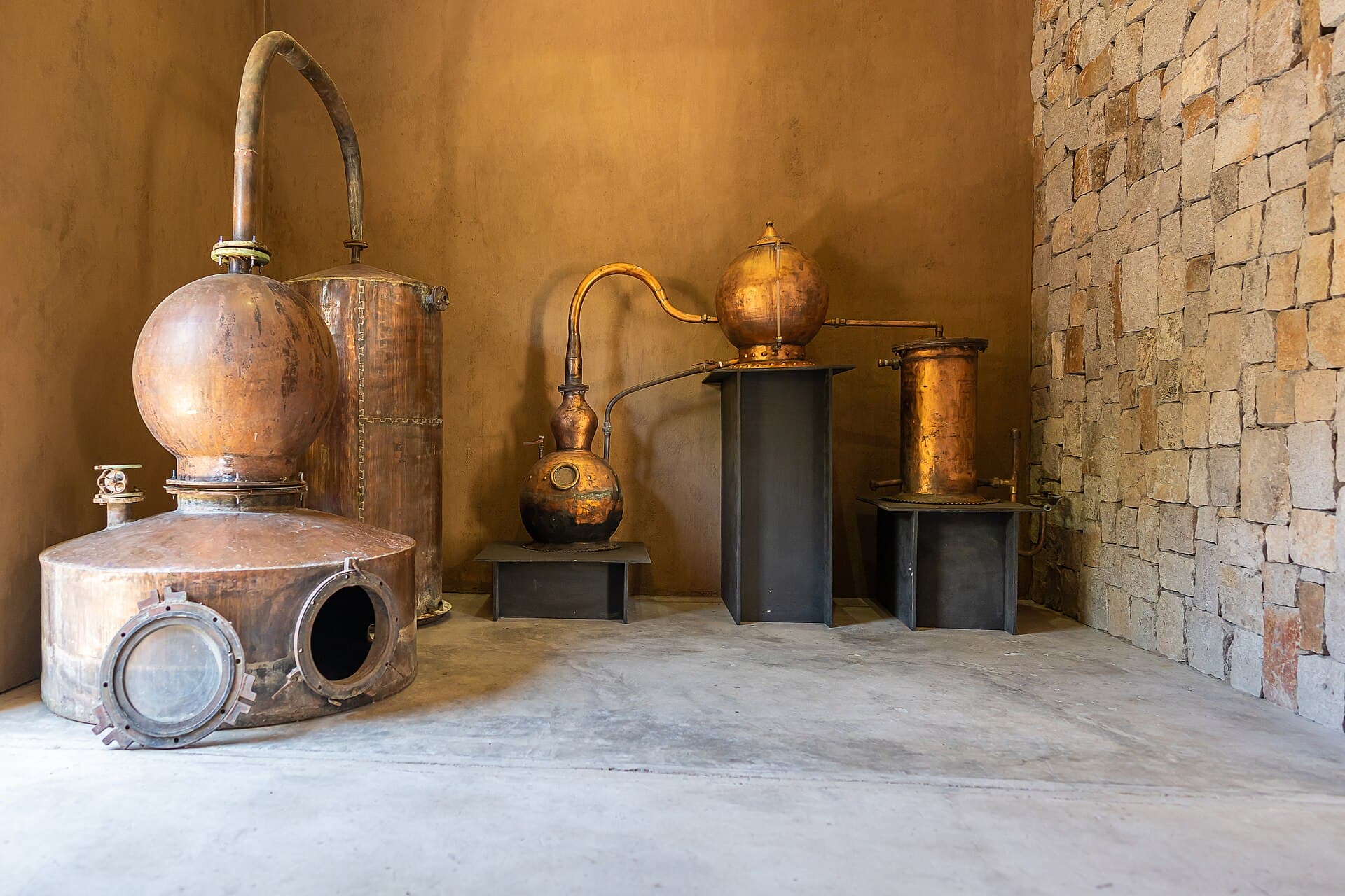 Alambiques de cobre em uma sala com parede de pedra, usados na destilação da cachaça