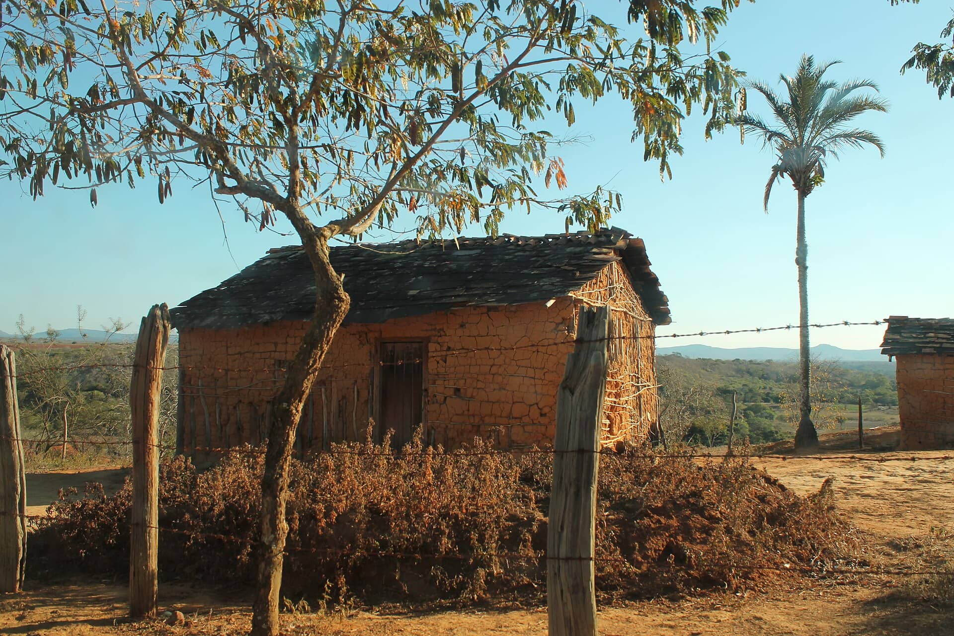 Casa de pau a pique de barro avermelhado, com telhado de telhas e cerca de arame, no interior do Brasil
