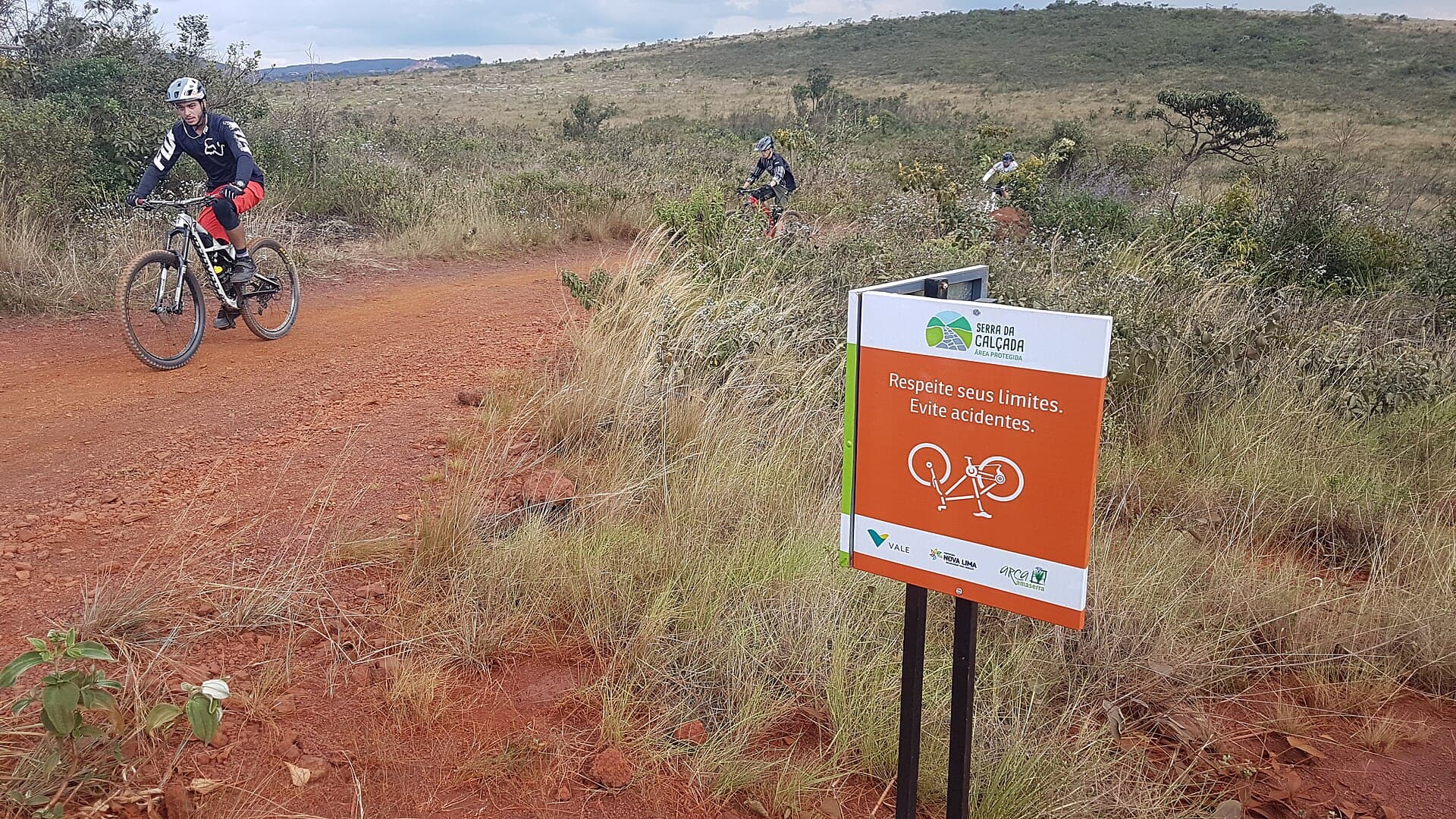 Ciclistas em trilha de terra vermelha ao lado de uma placa que pede respeito aos limites, na Serra da Calçada, em Minas Gerais
