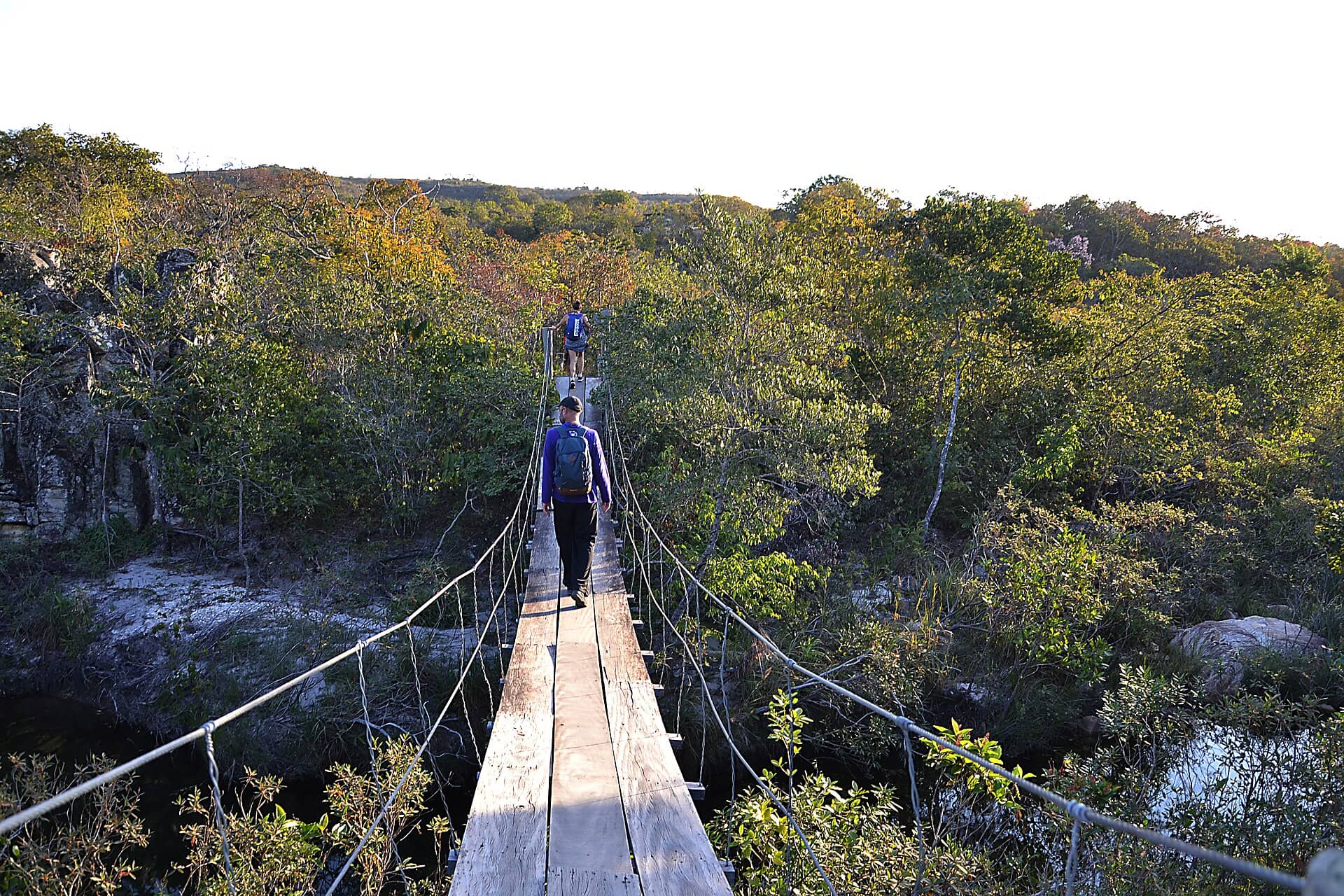 Dois caminhantes atravessam uma ponte pênsil de madeira em meio à vegetação de cerrado, na Serra da Canastra