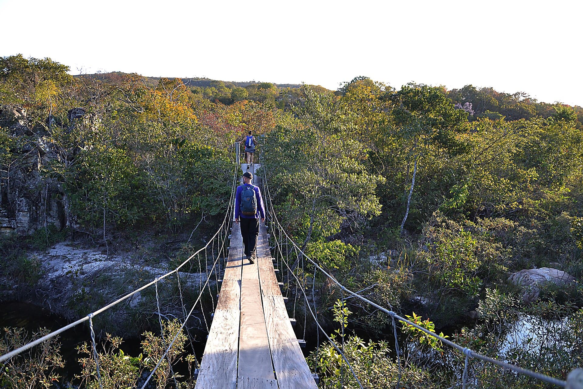 Dois caminhantes atravessam uma ponte pênsil de madeira em meio à vegetação de cerrado, na Serra da Canastra