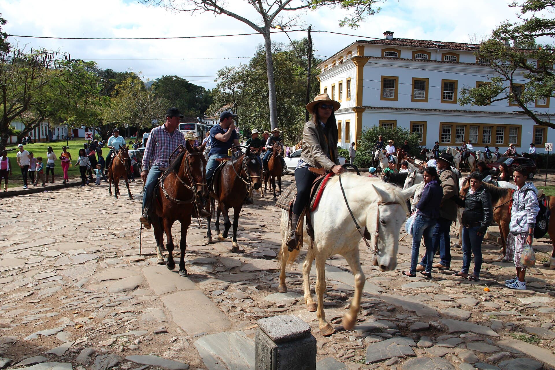 Grupo de cavaleiros com cavalos marrons e brancos passa por uma rua de pedras em Tiradentes, Minas Gerais