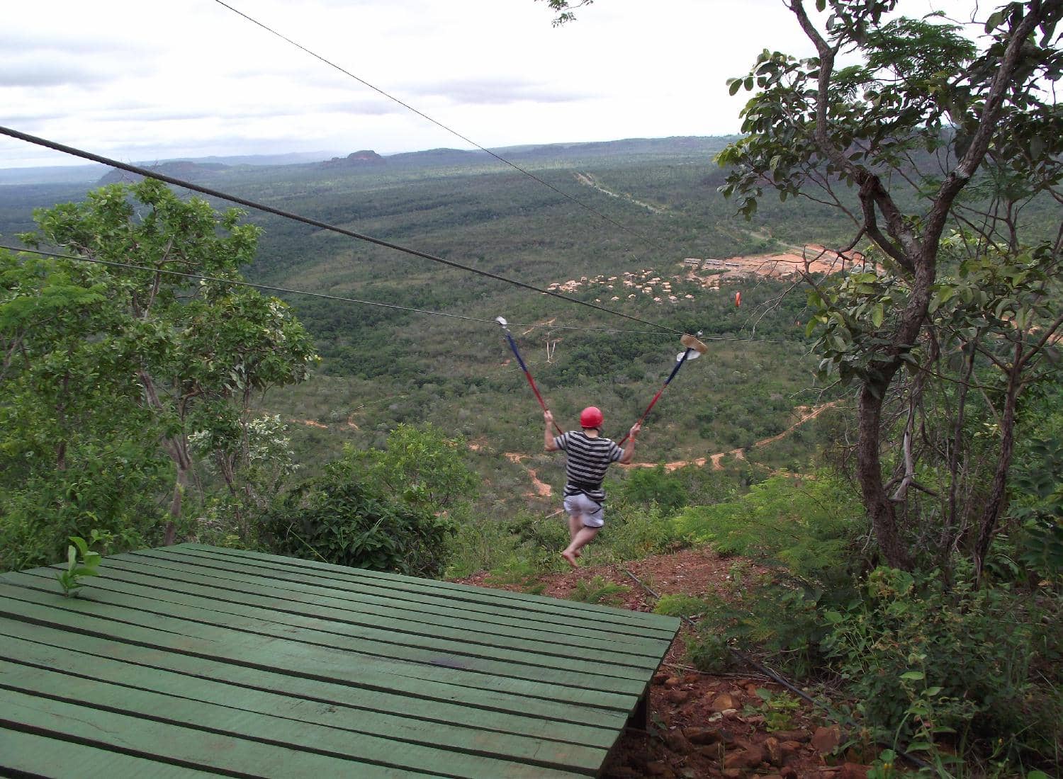 Praticante de tirolesa, de capacete, deixa uma plataforma de madeira e desce sobre um vale de cerrado