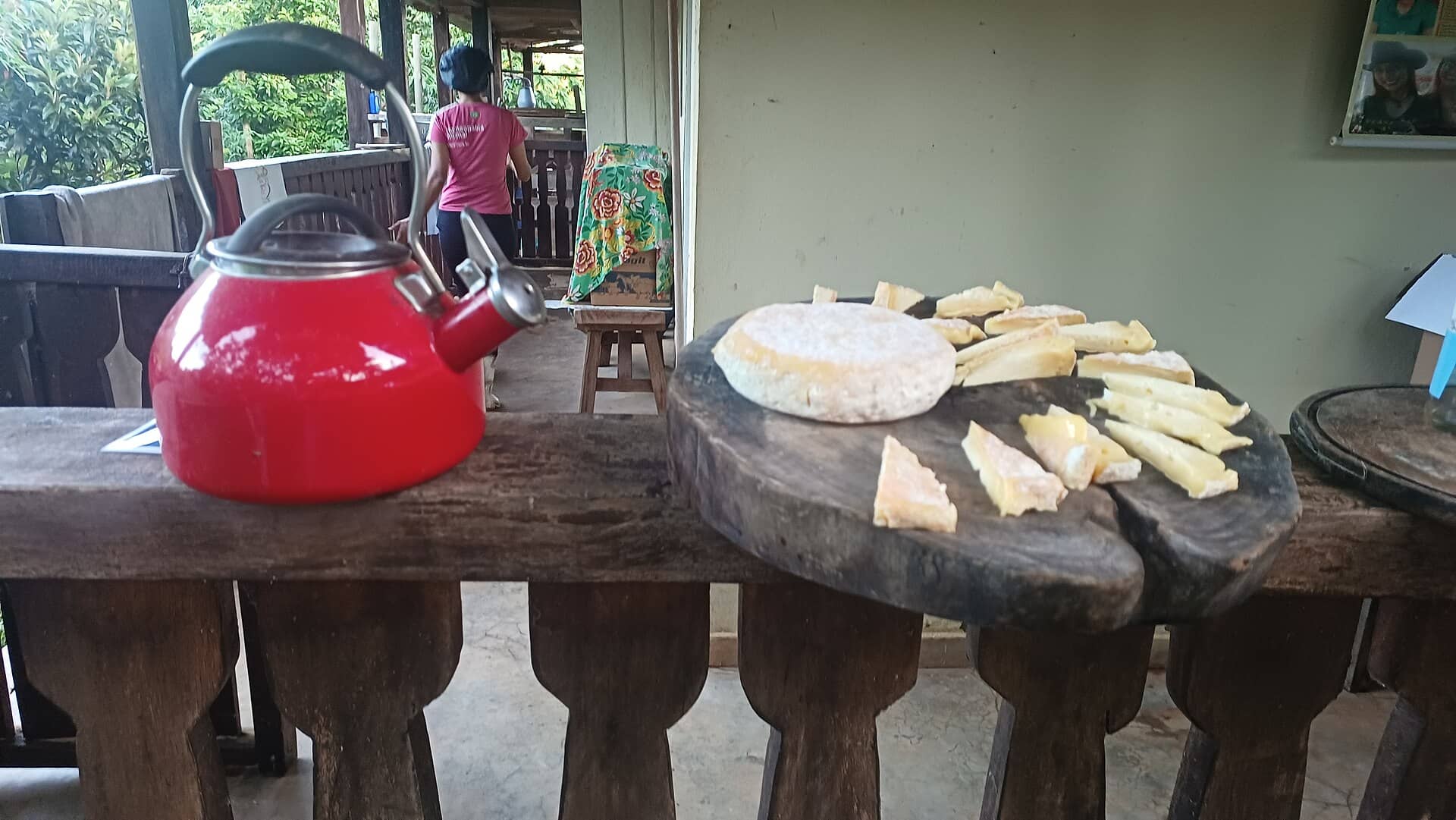 Queijo artesanal fatiado em uma tábua de madeira ao lado de uma chaleira vermelha, em varanda rústica