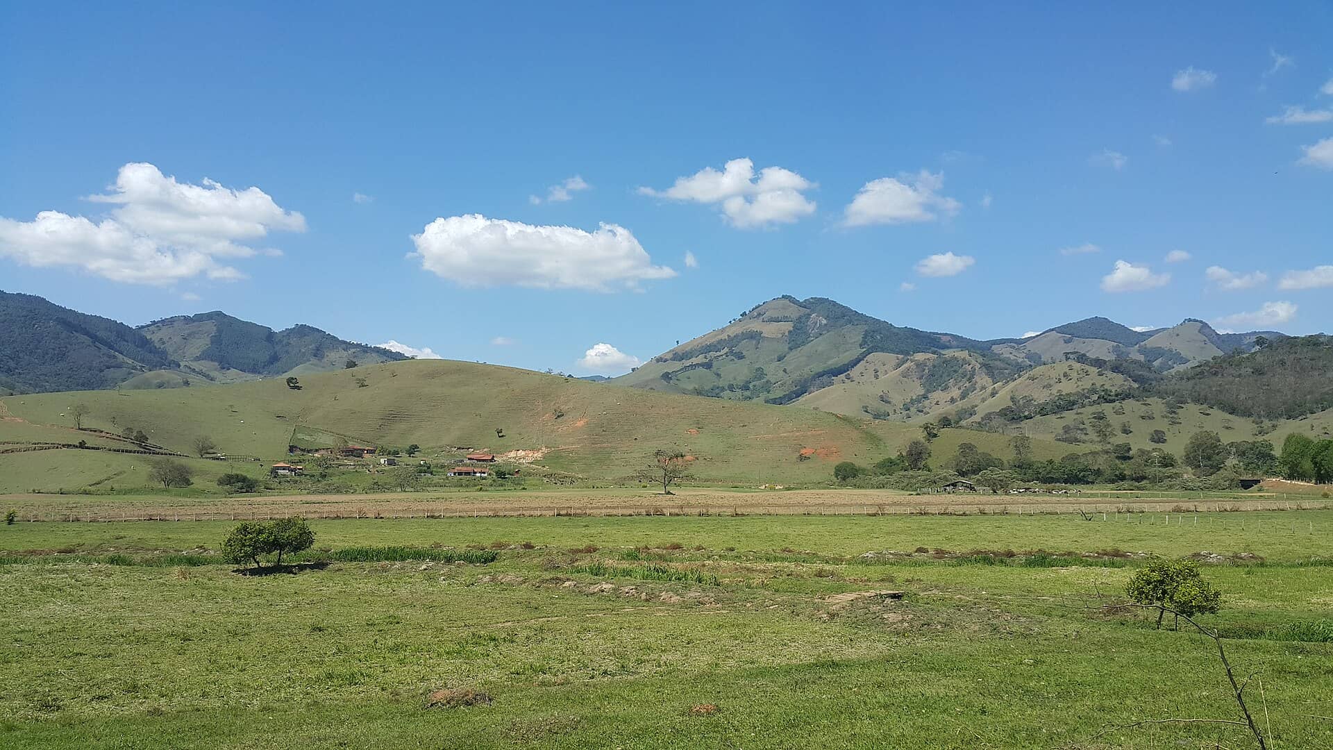 Vale de pastagens verdes cercado por morros sob céu azul, em Gonçalves, na Serra da Mantiqueira