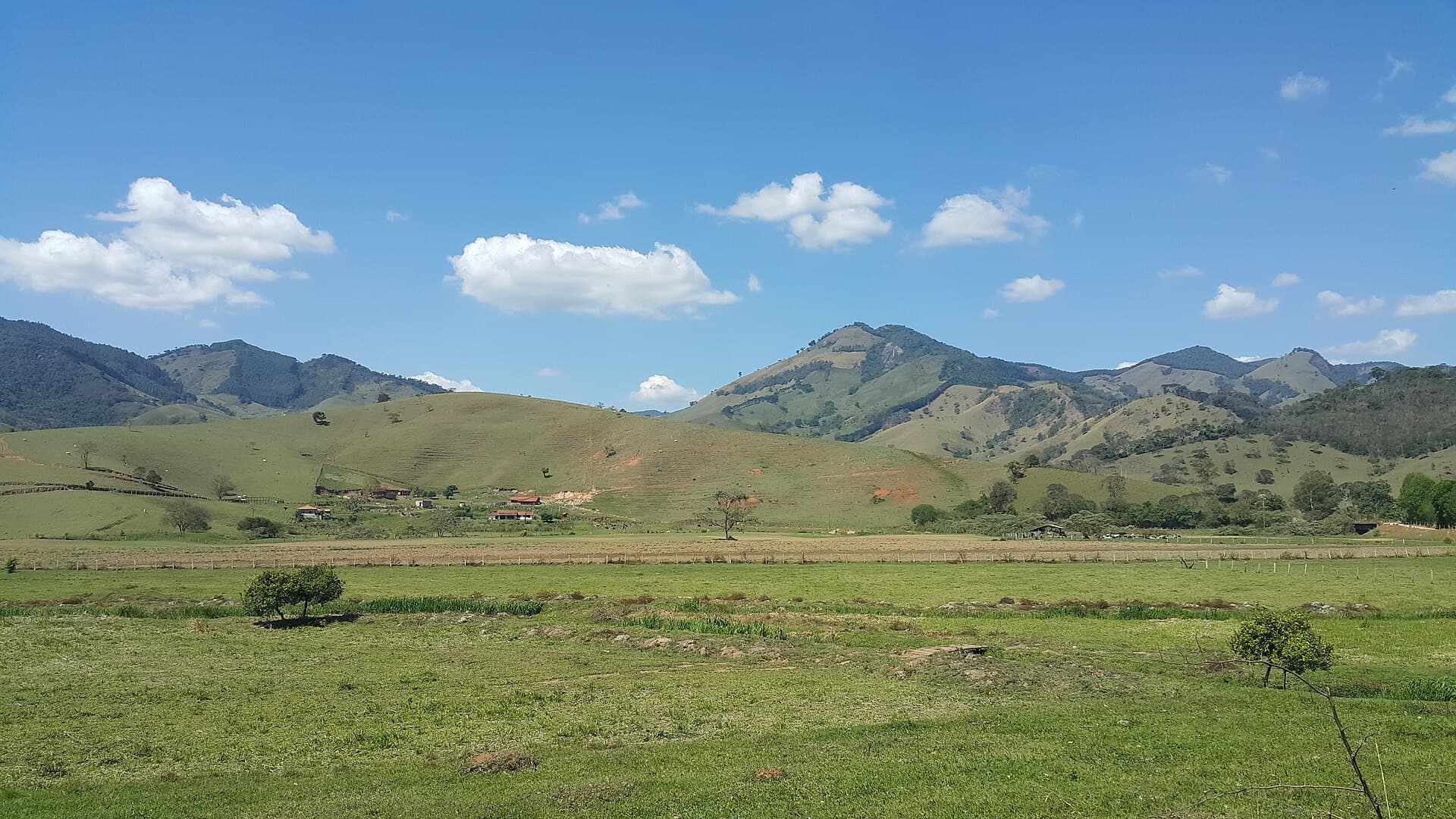 Vale de pastagens verdes cercado por morros sob céu azul, em Gonçalves, na Serra da Mantiqueira